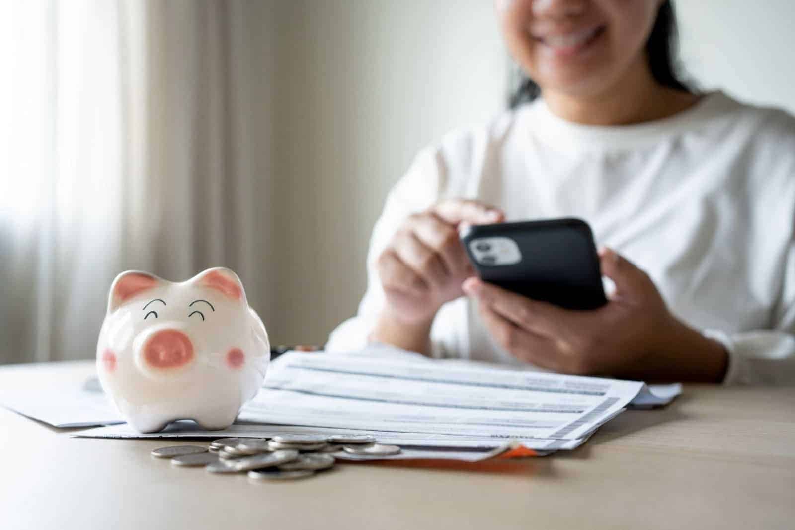 Lady seated at a table with a phone in hand and a piggy bank, coins, and paperwork on the table